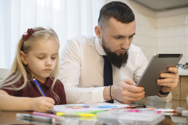 父亲爸爸上班前看着厨房里的平板电脑屏幕 而他的小女儿正在画画在线孩子社交媒体