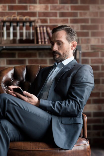 架子通信设备互动很好正经的帅哥坐在扶手椅上 一边拿着手机一边用独自现代西装