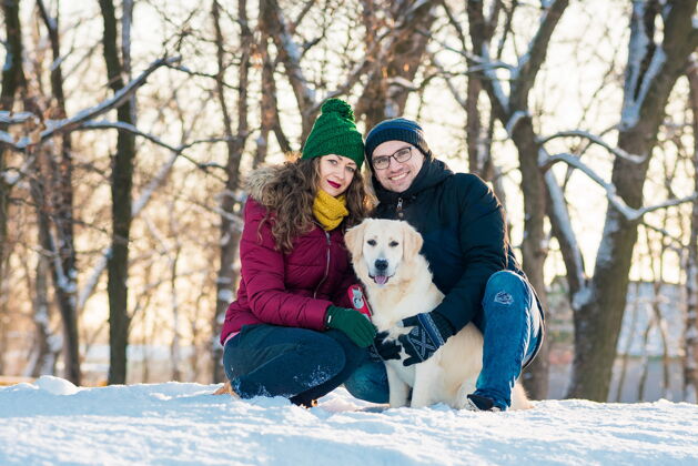 季节在阳光明媚的日子里 一对可爱的年轻夫妇和他们的金毛猎犬在冬季公园里玩得很开心微笑女人在冬天的雪乡 男人抱着狗欢乐人女人