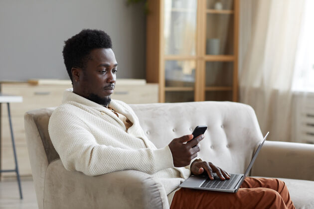 无线技术一个非洲男人一边在房间沙发上用笔记本电脑上网一边用手机读信息微笑室内成人
