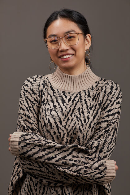 开衫毛衣一幅亚洲年轻女子的画像 她穿着优雅的衣服 戴着眼镜 微笑着站在灰色的墙上看面部表情积极的情绪