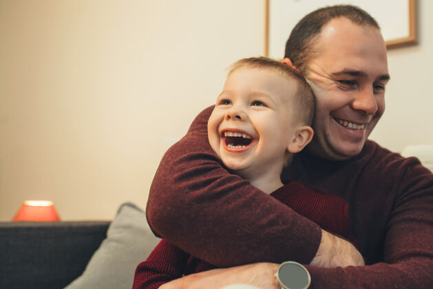 成人一个可爱的小孩笑着看着他的父亲拥抱他坐在家里的沙发上世代享受男孩