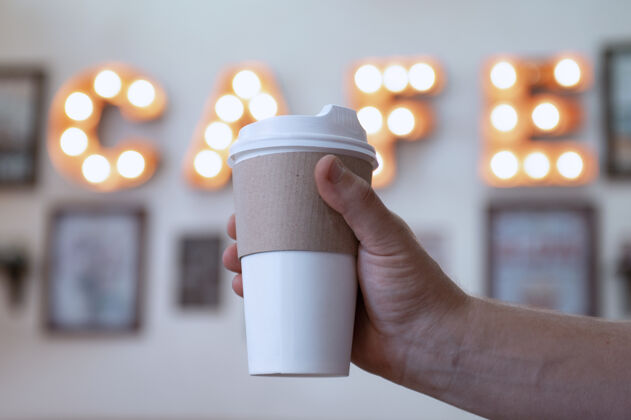 人一杯咖啡在咖啡馆里一个小伙子手里映衬着一片夜光招牌.mock-一个纸板环保马克杯年份早晨牛奶