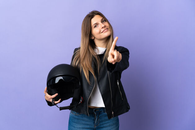 夹克年轻的斯洛伐克妇女拿着一个摩托车头盔 在紫色的背景上展示和举起一个手指摩托车手人金发