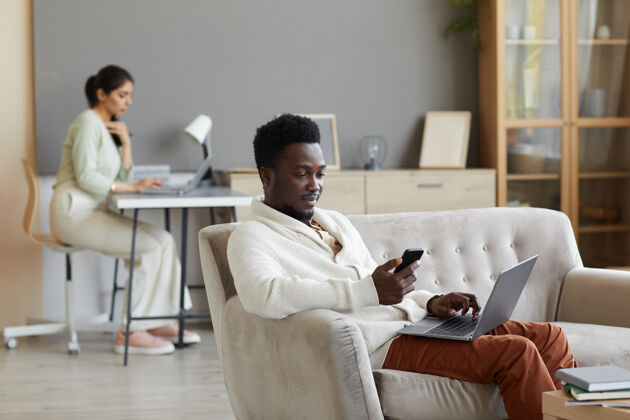 快乐坐在沙发上拿着笔记本电脑和手机的非洲男人和在家里墙里工作的女人室内商务女性人员
