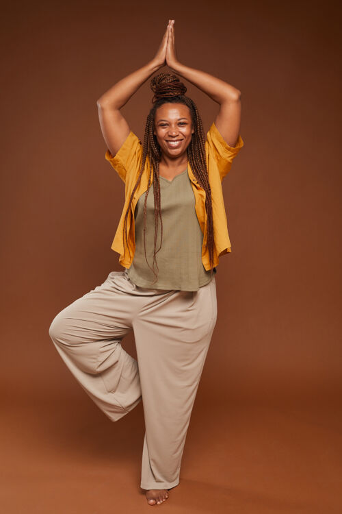 美女在棕色背景下 一位留着漂亮发型的非洲女人站在瑜伽姿势 对着镜头微笑享受自信健康