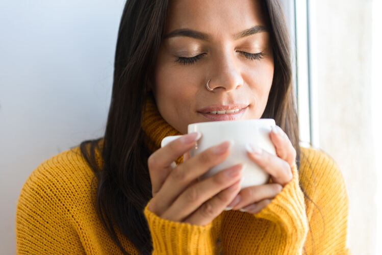 享受一个穿着毛衣的可爱的年轻女子坐在室内的窗边 喝着一杯茶坐着窗户茶