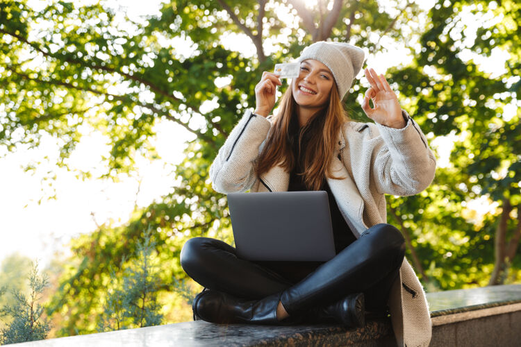 休闲快乐的年轻女子穿着秋大衣 戴着秋帽坐在户外 用笔记本电脑 展示塑料信用卡秋天学生年轻