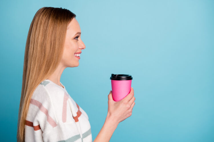 年轻开朗积极的女友的侧面照片 微笑着 望着空荡荡的空间 手里拿着一杯茶 隔离的淡蓝色墙壁咖啡套头衫时尚