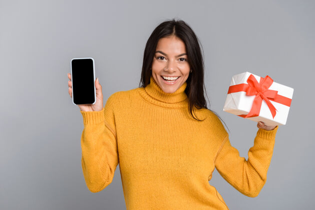 电话一个快乐随意的年轻女子的肖像被隔离在灰色的墙上 显示手机屏幕空白 手持礼品盒成人手机屏幕