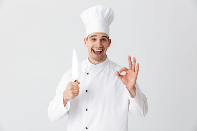 工作服快乐厨师穿着厨师制服拿着刀隔着白墙 行手势可爱吸引力Ok