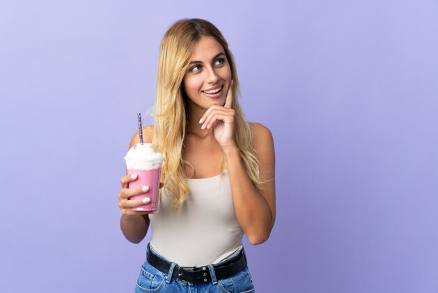 年轻人一个年轻的金发乌拉圭女人拿着草莓奶昔孤立地站在紫色地上 一边抬头一边思考着一个想法水果奶昔饮料