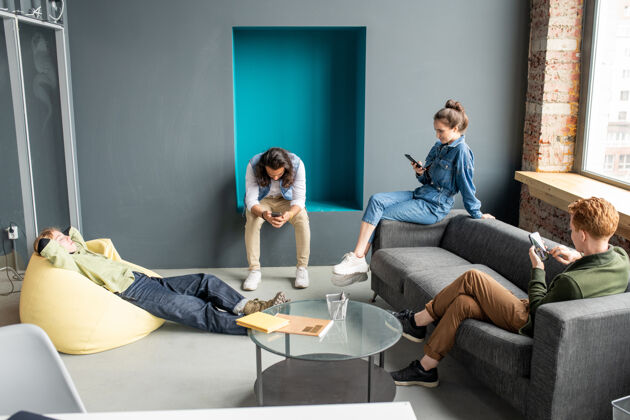 数字平板电脑三个年轻的安静的设计师或移动应用程序开发人员在智能手机上滚动 而金发女人躺在黄色的扶手椅上笔记本电脑讨论室内