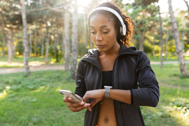 锻炼20多岁的年轻女子穿着黑色运动服 戴着耳机 在绿色公园里散步时看着手表女性精力充沛运动
