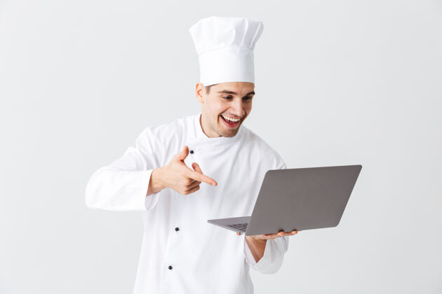 指向开朗的厨师穿着制服站在白色的墙上 手里拿着笔记本电脑美食家看笔记本电脑