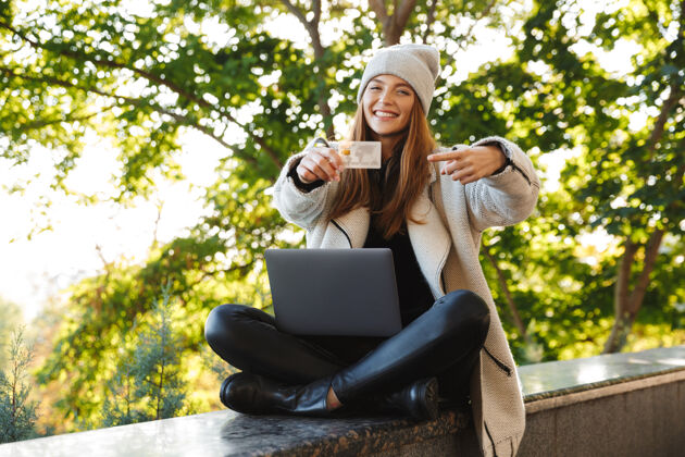信贷快乐的年轻女子穿着秋大衣 戴着秋帽坐在户外 用笔记本电脑 展示塑料信用卡年轻户外学生