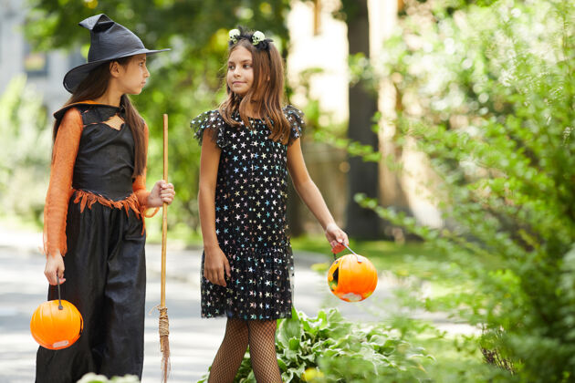 庆祝两个穿着万圣节服装的小女孩手里拿着南瓜形状的袋子 她们去户外享受美食玩耍孩子日子