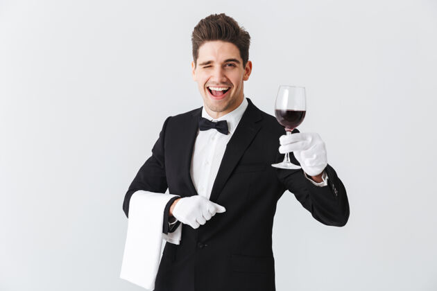 职业年轻帅气的侍者穿着燕尾服 端着一杯红酒隔着灰色的墙男人服务员杯子
