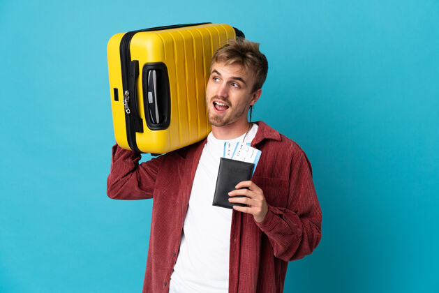 年轻年轻英俊的金发男人带着手提箱和护照在度假时被隔离在蓝色背景下人帅哥包