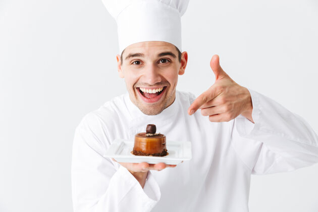 骄傲开朗的厨师穿着制服 在白色的墙上展示一个盘子上的糕点糕点男人盘子