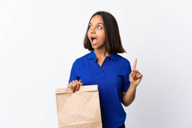 杂货店一个年轻的拉丁女人拿着一个杂货店的购物袋 孤立地站在白色的地板上思考着一个想法 手指向上超市想法看