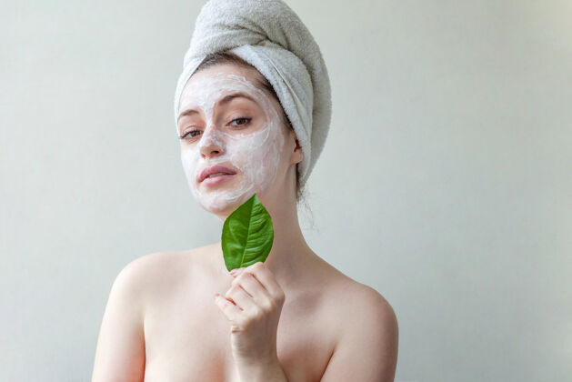 健康头戴毛巾 脸上涂有白色滋养面膜或乳霜 手拿绿叶 白色背景的美女肖像过程面膜面霜