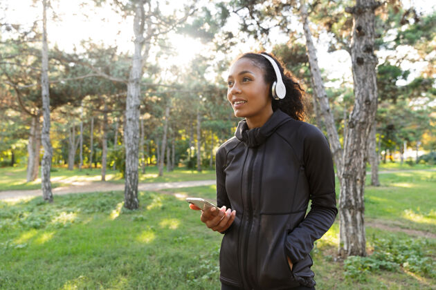音乐一位20多岁的健康女性穿着黑色运动服 戴着耳机 一边在绿色公园散步 一边用手机拍照女性耳机精力充沛