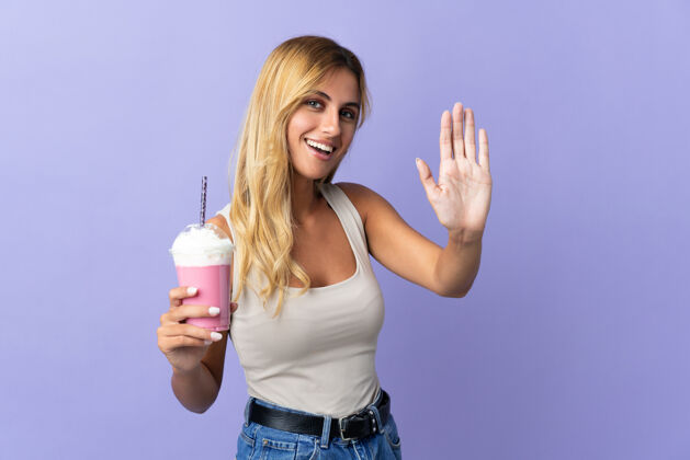 奶昔年轻的金发乌拉圭女人用草莓奶昔隔离在紫色的手与快乐的表情敬礼杯子金发年轻人