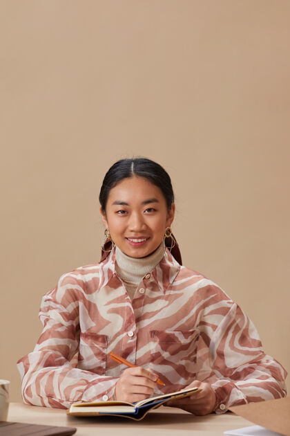 休闲服一幅年轻的亚洲女子坐在桌旁 在笔记本上写字时微笑的画像人大学生桌子