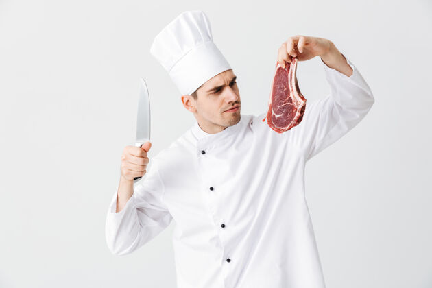 持有开朗的厨师穿着制服展示生牛排拿着刀隔着白墙薯条厨房餐