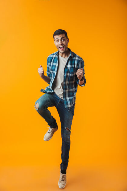 自信一个穿着格子衬衫的快乐的年轻人的全长肖像被隔离在橙色的墙上 跳跃庆祝成功男时尚肖像