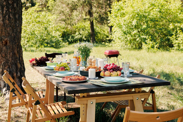 正式花园在阳光明媚的日子里 在松树下 一堆椅子围绕着一张木制的节日餐桌 桌上摆满了自制的食物和饮料 新鲜的水果和鲜花野餐桌夏天餐