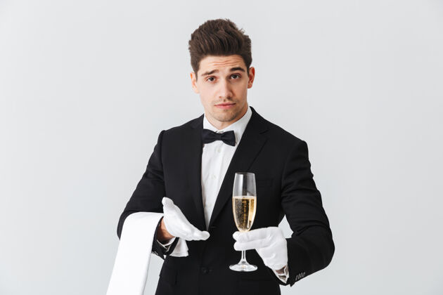 饮料身着燕尾服的微笑年轻侍者的画像在白墙上为您奉上一杯香槟服务员人杯子