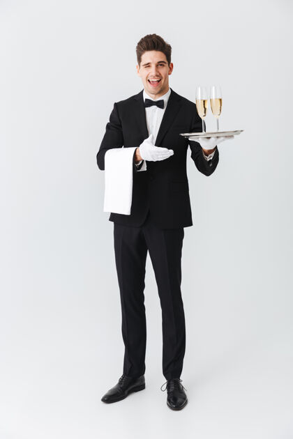 正式服装一位身着燕尾服 手持托盘 满脸笑容的年轻侍者的全长肖像 白色墙壁上有两杯香槟液体男性服务