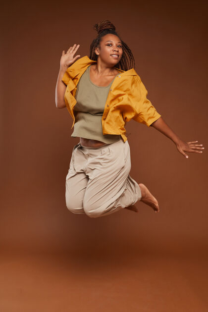 舞蹈穿着时尚服装的非洲快乐女人在棕色背景下跳跃的肖像艺术和娱乐感官平衡