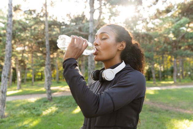 自然20多岁的女运动员穿着黑色运动服 戴着耳机 一边喝水一边穿过绿色公园运动非洲保健
