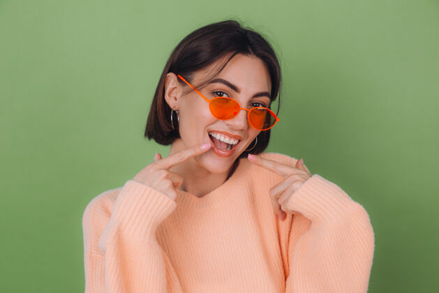 女孩年轻时尚的女人穿着休闲的桃色毛衣 戴着橙色眼镜 隔着绿橄榄色的墙壁 积极地微笑着 用食指指着白色的牙齿 复制空间牙医脸牙齿