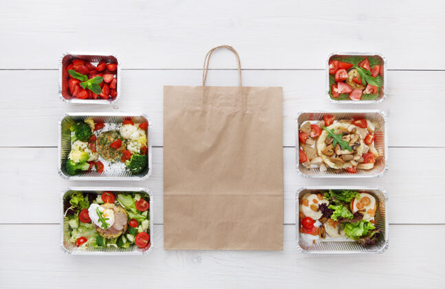 健康健康食品配送 日常膳食和零食营养 蔬菜 肉类和水果在铝箔盒和牛皮纸包装俯视图 平放在白色木材与复印空间午餐早餐蔬菜