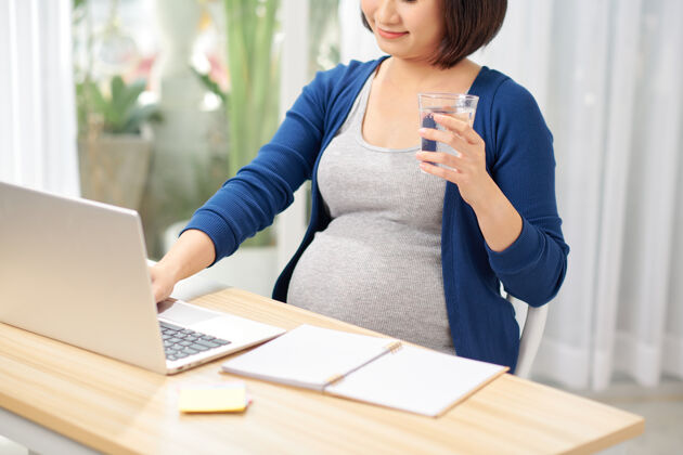 电脑孕妇在家办公桌旁拿着电脑和饮用水亚洲怀孕家庭办公室