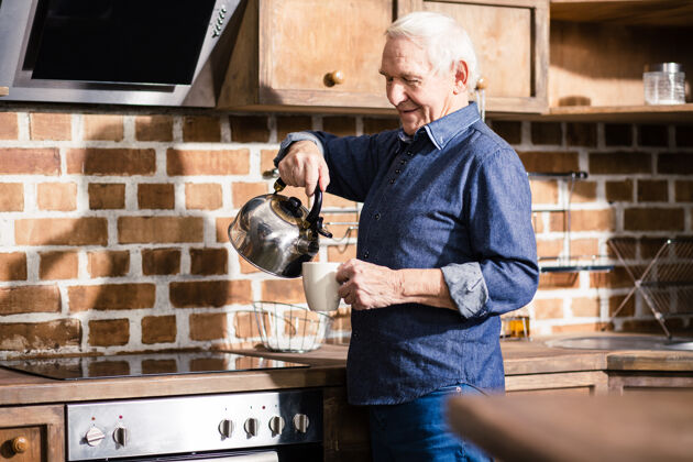 父母快乐的老人站在厨房里从水壶里倒茶早晨老年人厨房