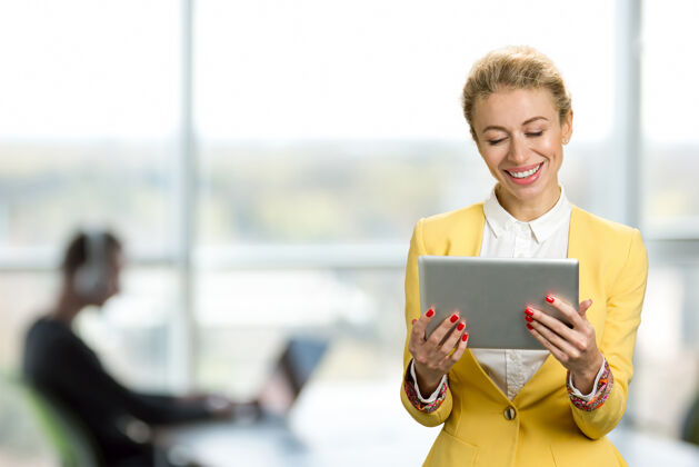 平板电脑面带微笑的商务女士拿着电脑平板电脑开朗的年轻商务女士看着电脑平板电脑 微笑着站在办公窗口公司工作成功