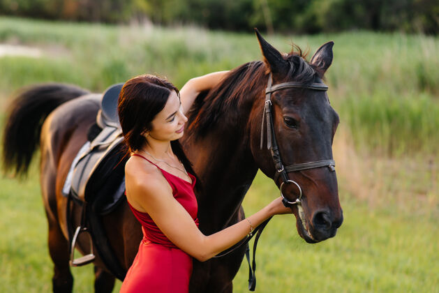 马厩日落时分 一个穿着红裙子的年轻漂亮女孩和一匹纯种种马在牧场上合影热爱和爱护动物年轻女骑师活动