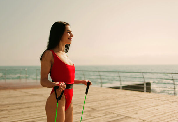肌肉穿着红色运动紧身衣的年轻女性在沙滩上用扩张器做有氧运动橡胶运动健身