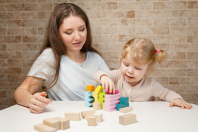 家庭可爱的小女孩和年轻的妈妈在桌子上玩积木木制玩具学习游戏房子