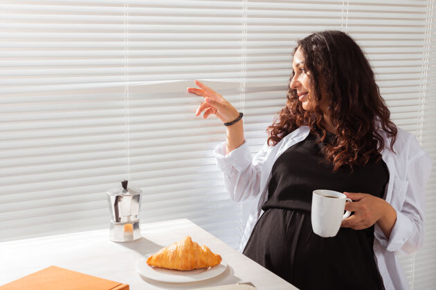 腹部快乐怀孕的年轻漂亮女人透过百叶窗看在她的早餐与咖啡专业知识黑发责任
