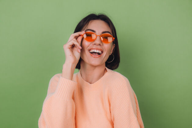 时尚年轻时尚的女士穿着休闲的桃色毛衣 戴着橙色的眼镜 隔着绿橄榄色的墙壁 开心的 积极的笑着 围着复制空间转魅力眼镜年轻