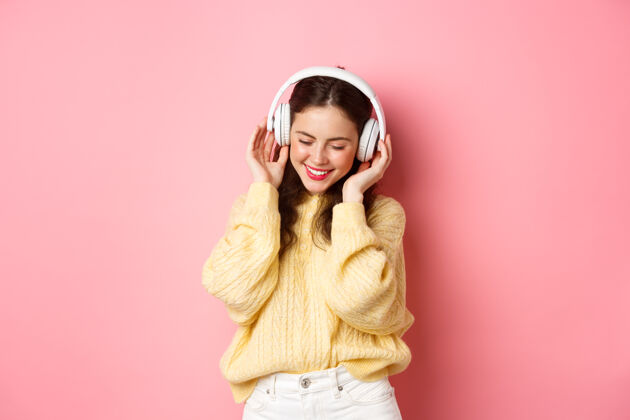 欢呼迷人的黑发女孩的肖像欣赏音乐 跳舞和微笑高兴 站在粉红色的墙壁复制空间魅力迷人耳机