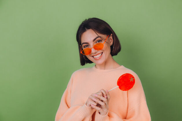 女士年轻时尚的女士穿着休闲的桃色毛衣 戴着橙色的眼镜 隔离在绿色橄榄色的墙上 带着橙色棒棒糖的正面微笑复制空间魅力积极漂亮