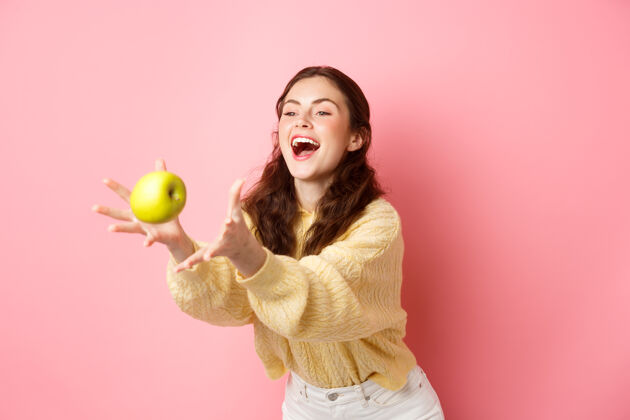 黑发快乐可爱的女孩抓到一个苹果 笑了起来 时间健康午餐充满水果 站在粉红色的墙壁快乐欢呼年轻