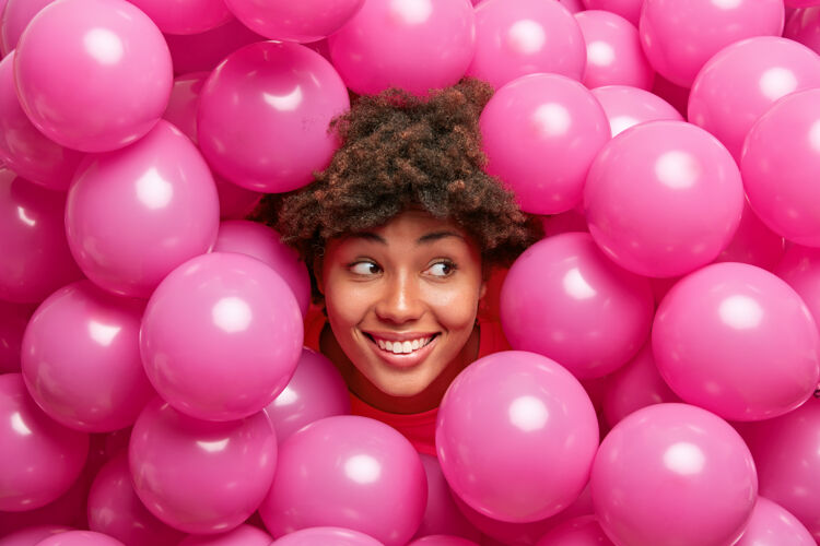 女性浓密卷发的女人笑容满面看起来很对 在充气的氦粉色气球周围摆出喜庆的姿势面部生日庆祝